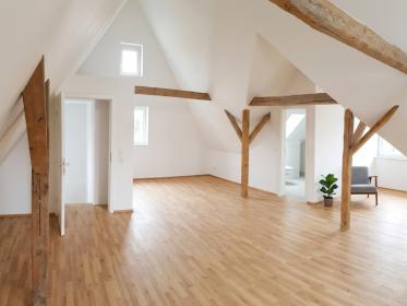 elles, modernisiertes Spitzboden-Zimmer mit freiliegenden Holzbalken und neu verlegtem Parkettboden; weiße Wände und eine minimal gestellte Armchair-Pflanzen-Ecke.