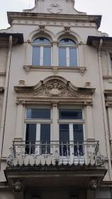 Historische Fassade in hellem Beige mit dekorativem Balkon und reich verzierten Fenstern, vor Renovierung leicht verschmutzt.