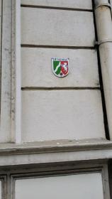 Nahaufnahme einer Außenwand mit kleinen Abplatzungen und dem Denkmal‑Schild, Oberfläche vor Ausbesserung.