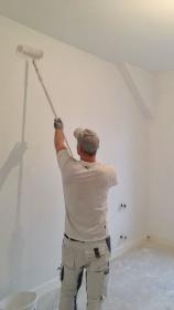 Maler arbeitet mit Farbroller an einer Zimmerwand, heller Raum im Renovierungszustand.