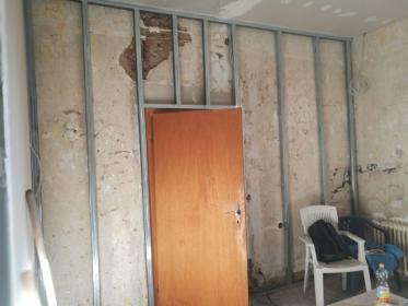 Innenaufnahme eines Raums im Rohbau: Metallständer, freiliegende Putzflächen, geöffnete Holztür und provisorische Stühle — Zustand vor Sanierung.